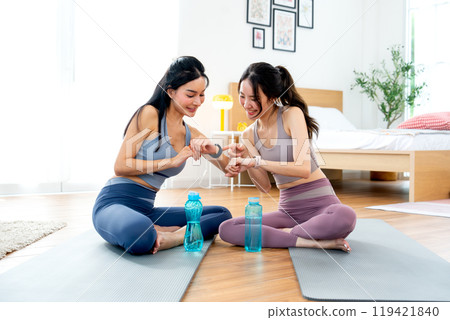 Side view two Asian women with exercise clothes sit on yoga mat and look and check their smart watch after workout in the house. 119421840