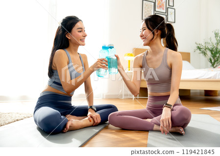 Two Asian women with exercise clothes sit on yoga mat and hold bottle of water then cheers together and relax after workout in the house. Two Asian women with exercise clothes sit on yoga mat and hold bottle of water then cheers together and relax after workout in the house. 119421845