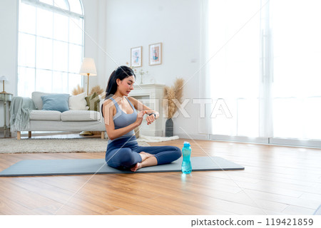 Pretty Asian woman with exercise clothes sit on mat in room with look at wristwatch during yoga exercise with happiness. 119421859