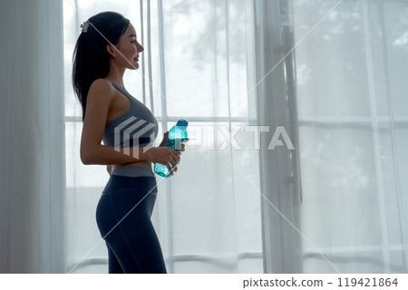 Pretty Asian woman with exercise clothes hold the bottle stand in front of curtain and glass door and also look outside with happiness. 119421864