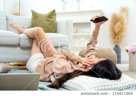 Beautiful young Asian woman lie on sofa the floor in living room of her house enjoy to using mobile phone with happiness. 119421869