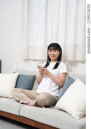 一位女士坐在客廳的沙發上操作智慧型手機 一位女士坐在客廳的沙發上操作智慧型手機 119422626