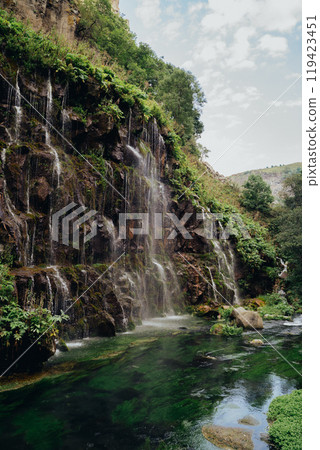A tranquil waterfall cascades down rugged cliffs surrounded by lush green foliage at noon 119423451
