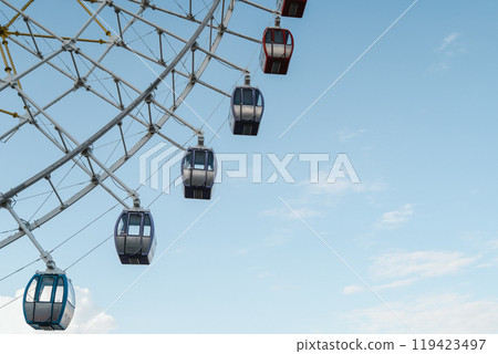 Part of a Ferris wheel spins in a sunny amusement park under a clear blue sky 119423497