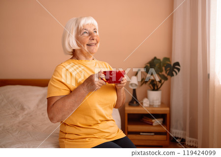 Caucasian blonde senior woman sitting on a white bed with a cup in her hand. Female relaxing and drinking cup of hot coffee or tea in the bedroom. 119424099