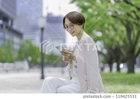 A businesswoman in an office district sitting on a bench and operating a smartphone 119425531