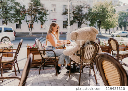 A woman sits cafe with a teddy bear next to her. The scene is set in a city with several chairs and tables around her. The woman is enjoying her time at the outdoor cafe. 119425810