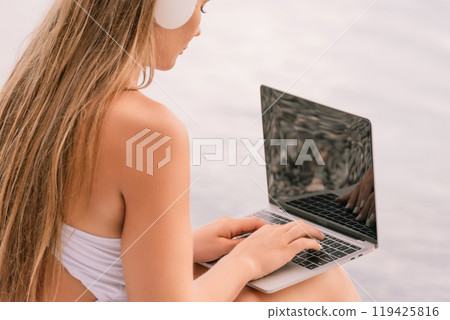 A woman is sitting on a beach with a laptop in her lap. She is wearing headphones and she is listening to music. 119425816