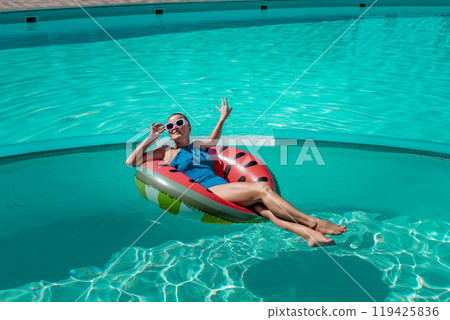 Happy woman in a swimsuit and sunglasses floating on an inflatable ring in the form of a watermelon, in the pool during summer holidays and vacations. Summer concept. Happy woman in a swimsuit and sunglasses floating on an inflatable ring in the form of a watermelon, in the pool during summer holidays and vacations. Summer concept. 119425836