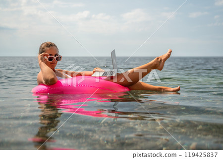 A woman is floating in a pink inflatable raft with a laptop in her hand. She is smiling and she is enjoying her time in the water. 119425837