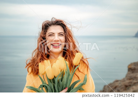 Portrait of a happy woman with hair flying in the wind against the backdrop of mountains and sea. Holding a bouquet of yellow tulips in her hands, wearing a yellow sweater 119425842