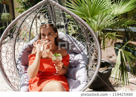 A woman is sitting in a hammock on a patio. The patio is surrounded by potted plants and has a few chairs and tables. The woman is drinking a beverage and she is enjoying her time. 119425980