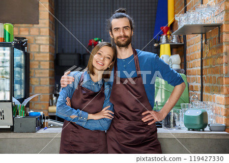 Hugging couple of colleagues partners young man and woman in aprons at workplace Hugging couple of colleagues partners young man and woman in aprons at workplace 119427330