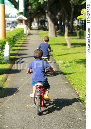 Two boys riding bicycles along a shaded pathway in a park. They are wearing matching blue shirts, with the trees and green lawn framing their journey. 119427486