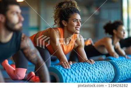 Energetic woman in orange top participating in group fitness class, using foam roller for exercise, showcasing enthusiasm and determination in gym setting 119428282