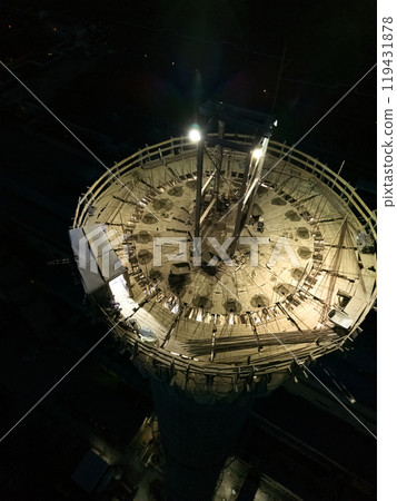 Vertical photo.Illuminated construction site at dusk.Dangerous work at night.Workers build a high chimney for a plant.Contractor repairs a chimney on a high-rise building.Worker in a hardhat on site 119431878