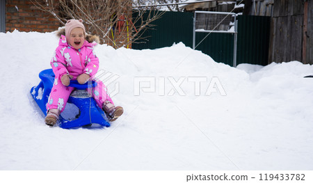 A child sleds down a snowy hill in the backyard, enjoying the winter. Copy Space. In a bright pink jumpsuit, a little girl sleds down a snow hill built by her parents. A fun winter day. 119433782