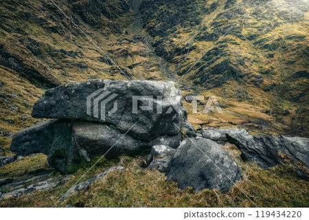 A large rock sits atop a grassy slope, surrounded by a rugged valley. The landscape features a mix of dry grass and rocky terrain under a cloudy sky. A large rock sits atop a grassy slope, surrounded by a rugged valley. The landscape features a mix of dry grass and rocky terrain under a cloudy sky. 119434220