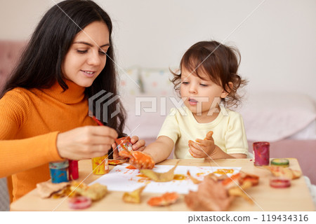 Cute little girl and mom spending good time painting hands and autumn yellow leaves together Cute little girl and mom spending good time painting hands and autumn yellow leaves together 119434316