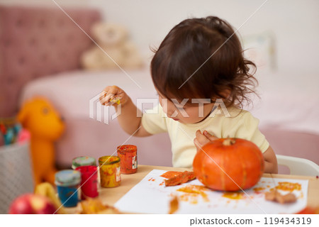 happy little child girl painting Halloween pumpkin. smiling baby girl drawing autumn pumpkin in yellow paint 119434319