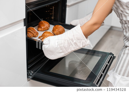 female baker taking tray with fresh croissants from oven in modern kitchen, close-up female baker taking tray with fresh croissants from oven in modern kitchen, close-up 119434328