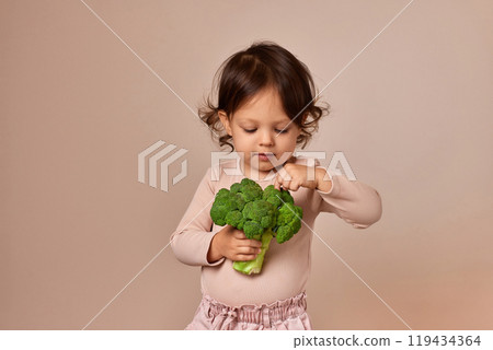 smiling child girl holding fresh green raw broccoli on beige background. healthy baby food. kid loves vegetables smiling child girl holding fresh green raw broccoli on beige background. healthy baby food. kid loves vegetables 119434364