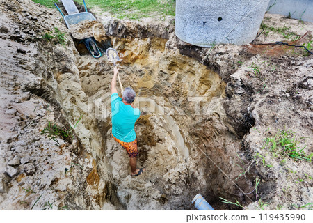 Homeowner manually digs excavation for an autonomous sewerage system in rural area using bayonet shovel. Homeowner manually digs excavation for an autonomous sewerage system in rural area using bayonet shovel. 119435990