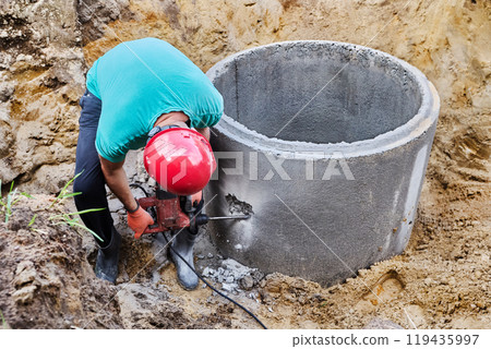 Using rotary hammer with chisel, worker punches hole for sewer pipe in concrete septic tank ring. 119435997