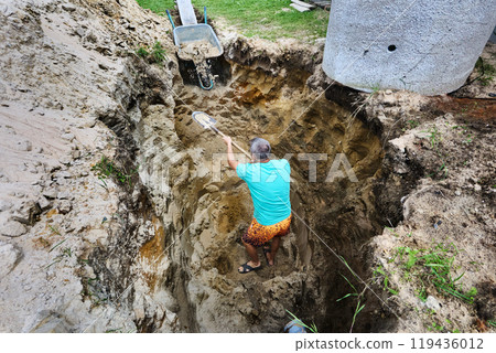 Homeowner throws soil into wheelbarrow while digging hole for septic tank. Homeowner throws soil into wheelbarrow while digging hole for septic tank. 119436012