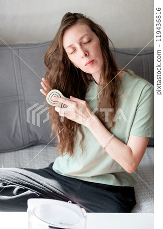 beautiful young woman with long wavy hair doing styling and combing it with a comb, looking in the mirror, sitting on the sofa, at home 119436816