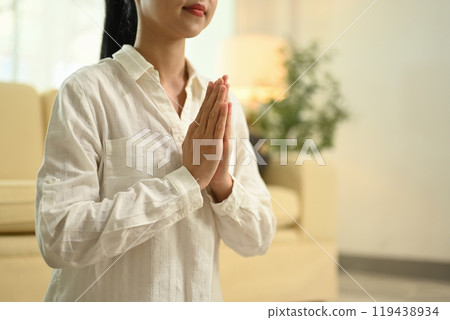 Calm woman doing yoga meditating in living room with her eyes closed and hands clasped 119438934
