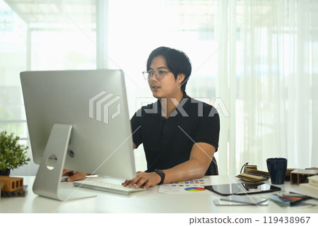Handsome asian architect looking at computer monitor working on project in his office Handsome asian architect looking at computer monitor working on project in his office 119438967