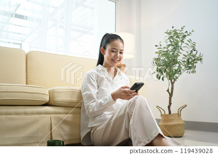 Beautiful young woman s sitting on floor at home and using mobile phone Beautiful young woman s sitting on floor at home and using mobile phone 119439028