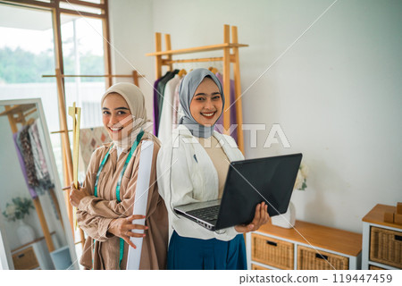 two female Muslim designers smile as they stand holding a laptop 119447459