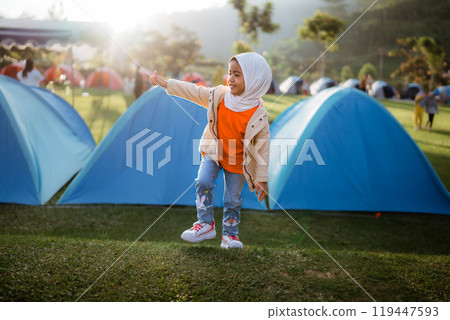 A little girl who is wearing a hijab is standing in front of a tent A little girl who is wearing a hijab is standing in front of a tent 119447593