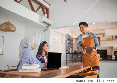two women work on laptops while ordering food from a male waiter two women work on laptops while ordering food from a male waiter 119447656
