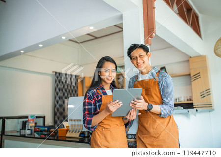 Indonesian man and woman in apron smiling using a tablet together Indonesian man and woman in apron smiling using a tablet together 119447710