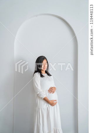 A pregnant woman in white dress stands by a wall, holding belly A pregnant woman in white dress stands by a wall, holding belly 119448113