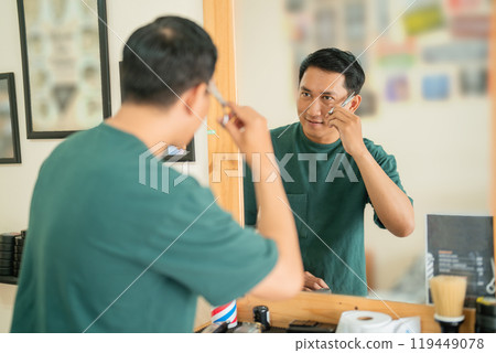 A man is carefully shaving his face while standing in front of a mirror 119449078