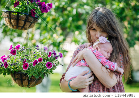 Young mother gently cradles her sleeping newborn baby girl amidst lovely hanging flower baskets in a lush garden 119449995