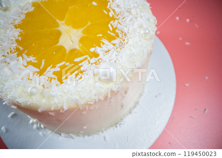 Delicious white cake decorated with orange and coconut flakes standing on pink background 119450023