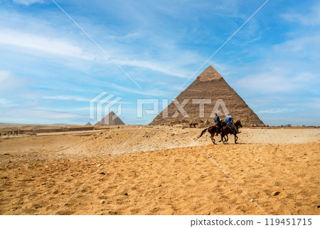 Two riders on black horses gallop against the background of the Pyramids of Khafren and Cheops in the sands of the Giza desert, Egypt Two riders on black horses gallop against the background of the Pyramids of Khafren and Cheops in the sands of the Giza desert, Egypt 119451715