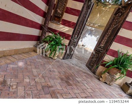 A courtyard with red and white walls, brick floor, potted plants, and a wooden door to another courtyard A courtyard with red and white walls, brick floor, potted plants, and a wooden door to another courtyard 119453244