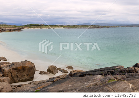 Evening at Gurteen Bay, Ireland, captures the peaceful shoreline with its clear turquoise waters and rocky formations. The soft white sand meets the sea under an overcast sky 119453758
