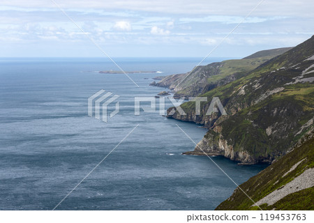 The dramatic cliffs of Slieve League in Ireland stretch along the Atlantic Ocean, with steep, rocky faces and verdant grassland. A small island with a lighthouse is visible offshore 119453763