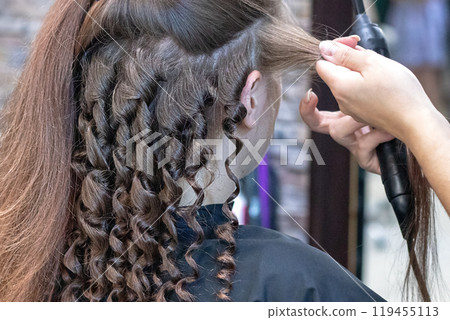 Hair stylist prepares a woman, makes a hairstyle for curls using a curling iron. Long brown natural hair 119455113