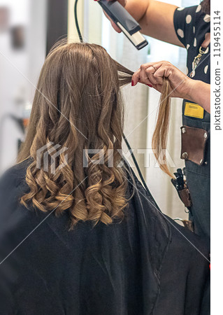 Hair stylist prepares woman makes curls hairstyle with curling iron. Long light brown natural hair Hair stylist prepares woman makes curls hairstyle with curling iron. Long light brown natural hair 119455114