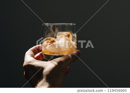 A glass filled with ice cubes and powdered Amanita muscaria mushroom on its surface, held in the hand, illuminated by natural sunlight. Preparing a homemade fly agaric elixir 119456005