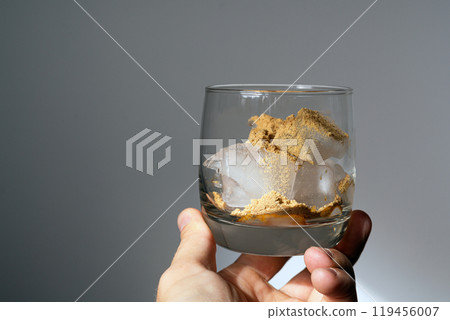 A glass filled with ice cubes and powdered Amanita muscaria mushroom on its surface, held in the hand, illuminated by natural sunlight. Preparing a homemade fly agaric elixir A glass filled with ice cubes and powdered Amanita muscaria mushroom on its surface, held in the hand, illuminated by natural sunlight. Preparing a homemade fly agaric elixir 119456007