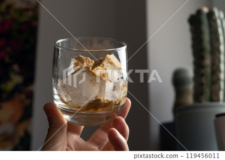 A glass filled with ice cubes and powdered Amanita muscaria mushroom on its surface, held in the hand, illuminated by natural sunlight. Preparing a homemade fly agaric elixir 119456011